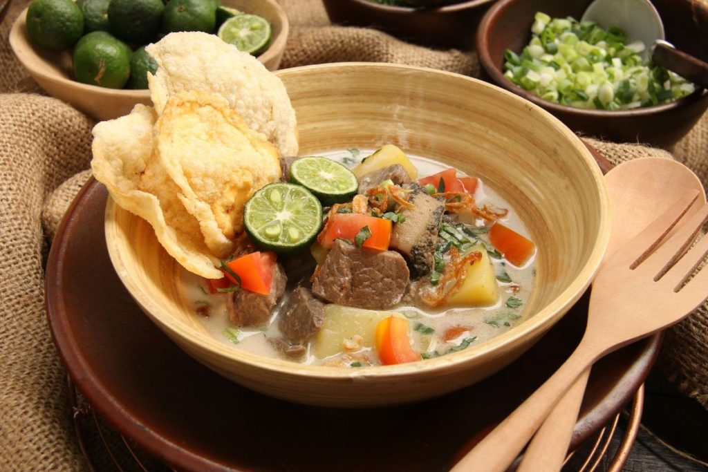 Semangkuk Soto Betawi kuah susu dengan potongan daging sapi empuk, kentang rebus, tomat, daun bawang, seledri, dan jeruk nipis sebagai pelengkap.